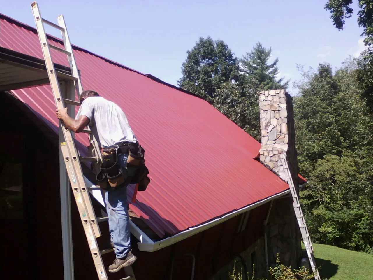 Roofer on a ladder beside a red metal roof and stone chimney — Soffit & Fascia Repair by Roofing Brothers in Caryville